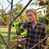 A man wearing a plaid shirt uses a cordless hedge trimmer in a garden, cutting a branch. The hedge trimmer is bright lime green and black, and the background shows a blurred green lawn and trees.